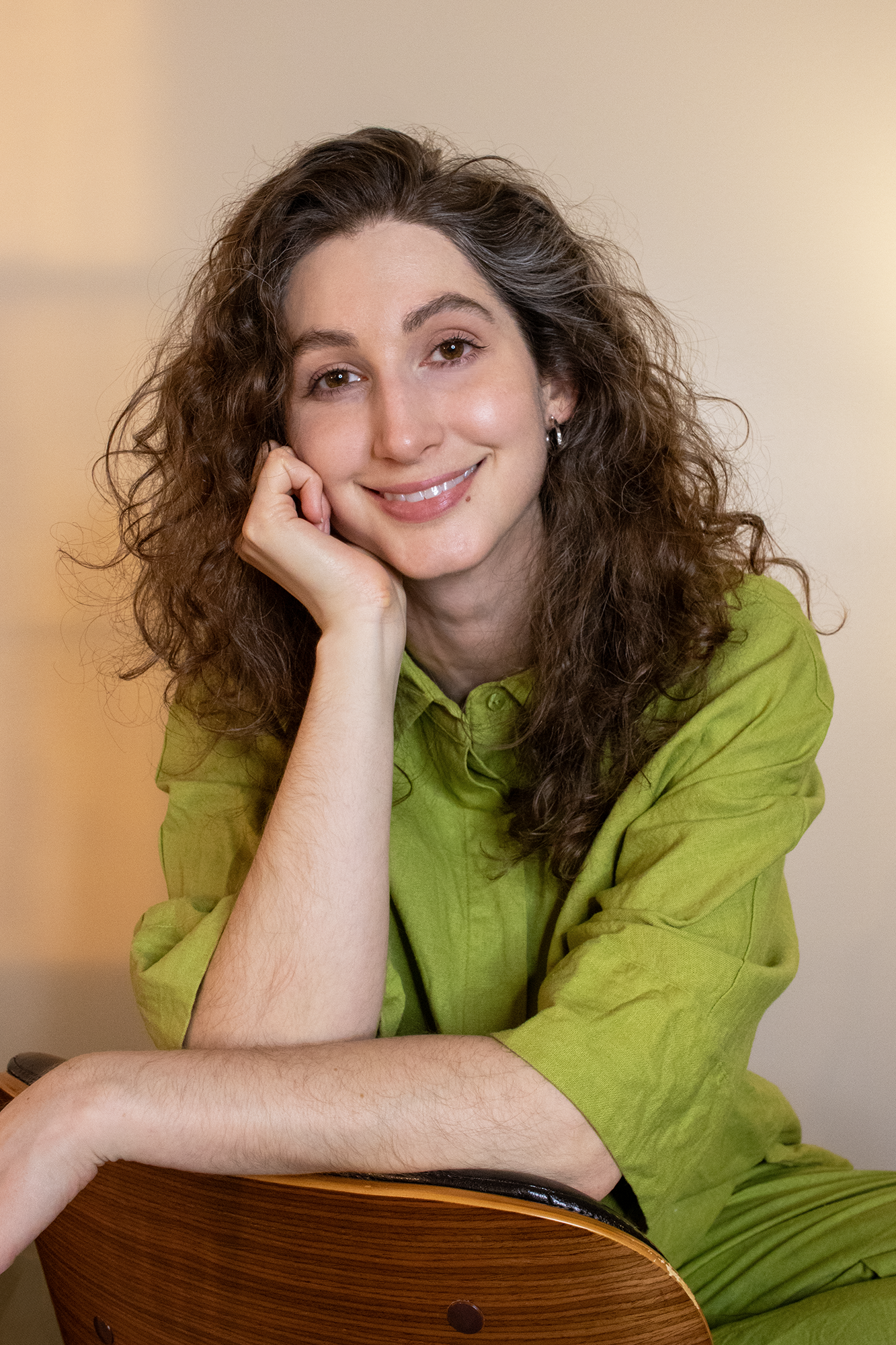 Geena Matuson, a white woman in her mid-30s with brown, curly hair just below shoulder length, wears a lime green, linen loose-fit top and pants. She is sitting on a chair backwards, her elbows draped over the back of a chair, with one arm raised to rest against her chin. Behind her is a blank, beige wall with a subtle hint of sunset light from an adjacent window.