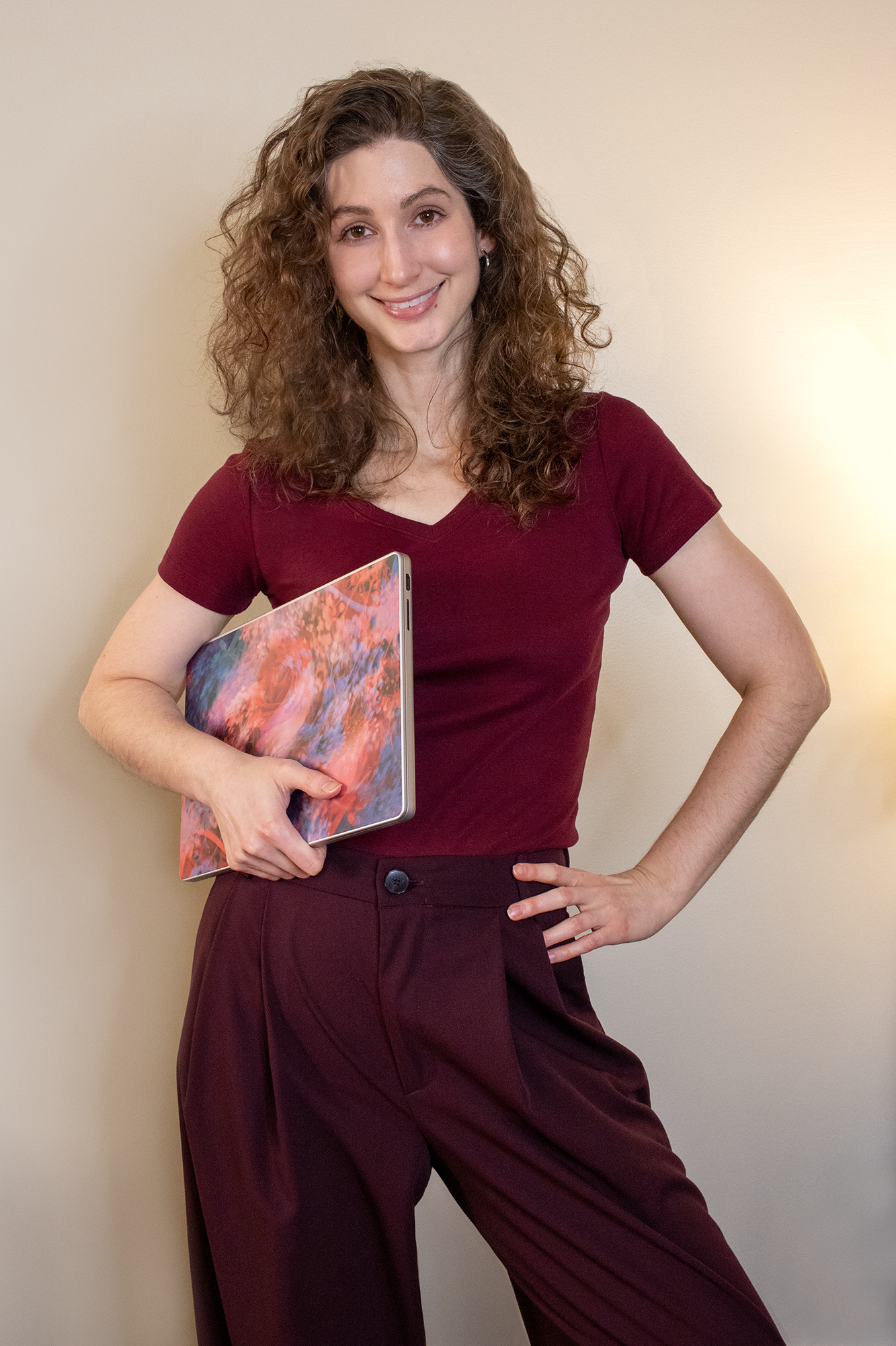 Geena Matuson, a white woman in her mid-30s with brown, curly hair just below shoulder length, wears a maroon fitted V-neck short-sleeve shirt and burgundy pants; image cropped at her knees. She has one hand on her hip, the other holding a laptop with its back to the camera. The laptop back has an abstract art print laptop skin, one of Geena's artworks.