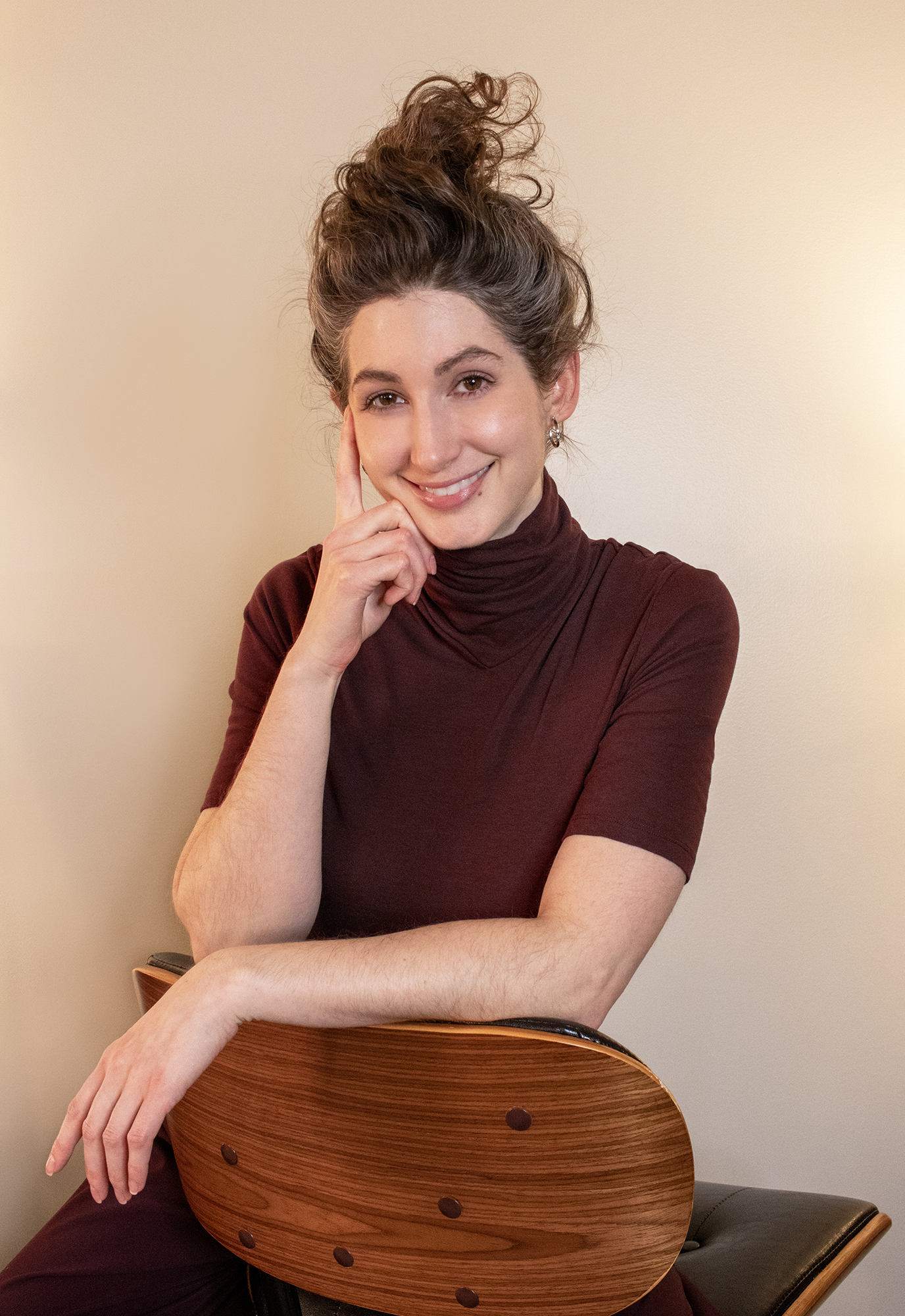 Geena Matuson, a white woman in her mid-30s with brown hair in a swoopy updo, wears a burgundy fitted turtleneck and smiles to the camera. She is sitting on a chair backwards, her elbows draped over the back of a chair, with one arm raised to rest against her chin. Behind her is a blank, beige wall.
